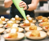 A person is piping whipped cream onto small cakes arranged neatly on plates. Each plate contains a cake and sliced strawberries, suggesting a dessert preparation setting. The scene is set in a professional kitchen with a focus on the dessert assembly process.