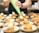 A person is piping whipped cream onto small cakes arranged neatly on plates. Each plate contains a cake and sliced strawberries, suggesting a dessert preparation setting. The scene is set in a professional kitchen with a focus on the dessert assembly process.