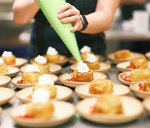 A person is piping whipped cream onto small cakes arranged neatly on plates. Each plate contains a cake and sliced strawberries, suggesting a dessert preparation setting. The scene is set in a professional kitchen with a focus on the dessert assembly process.