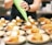 A person is piping whipped cream onto small cakes arranged neatly on plates. Each plate contains a cake and sliced strawberries, suggesting a dessert preparation setting. The scene is set in a professional kitchen with a focus on the dessert assembly process.