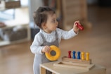 A toddler is wearing a grey romper and a white long-sleeve shirt. The child is standing by a wooden toy with pegs, holding a red peg in one hand and a yellow ring in the other. The background is softly blurred, indicating an indoor setting.