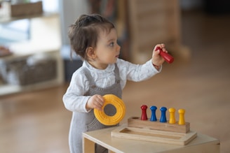 A toddler is wearing a grey romper and a white long-sleeve shirt. The child is standing by a wooden toy with pegs, holding a red peg in one hand and a yellow ring in the other. The background is softly blurred, indicating an indoor setting.