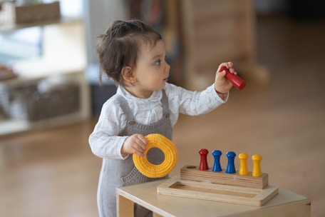 A toddler is wearing a grey romper and a white long-sleeve shirt. The child is standing by a wooden toy with pegs, holding a red peg in one hand and a yellow ring in the other. The background is softly blurred, indicating an indoor setting.