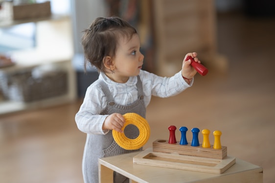 A toddler is wearing a grey romper and a white long-sleeve shirt. The child is standing by a wooden toy with pegs, holding a red peg in one hand and a yellow ring in the other. The background is softly blurred, indicating an indoor setting.