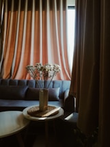 A cozy living room setting features a large, comfortable sofa in front of heavy, beige curtains that filter sunlight softly into the room. On a small, round table in the foreground, a white vase holds a bouquet of white flowers, placed on a woven mat.