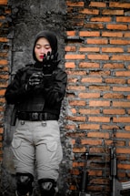 A person wearing a black tactical uniform with armor gloves stands in front of a brick wall with firearms resting against it. The individual looks focused and is adjusting their gloves.