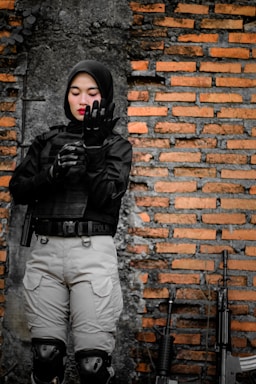 A person wearing a black tactical uniform with armor gloves stands in front of a brick wall with firearms resting against it. The individual looks focused and is adjusting their gloves.
