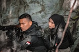 Two individuals dressed in tactical gear are positioned in a dense, foliage-rich environment. One person is aiming a rifle while the other appears to be communicating or signaling behind them. Both wear dark clothing and have Indonesian flags on their uniforms.