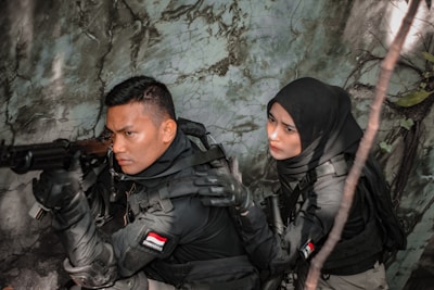 Two individuals dressed in tactical gear are positioned in a dense, foliage-rich environment. One person is aiming a rifle while the other appears to be communicating or signaling behind them. Both wear dark clothing and have Indonesian flags on their uniforms.