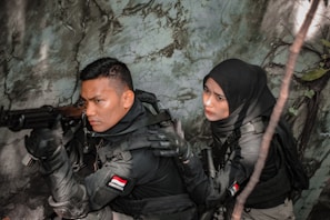 Two individuals dressed in tactical gear are positioned in a dense, foliage-rich environment. One person is aiming a rifle while the other appears to be communicating or signaling behind them. Both wear dark clothing and have Indonesian flags on their uniforms.