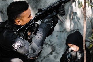 A person dressed in tactical gear aims a rifle upward while another person in a black hood is partially visible in the background. The setting appears to be an outdoor area with some foliage.