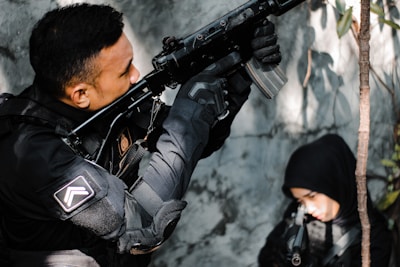 A person dressed in tactical gear aims a rifle upward while another person in a black hood is partially visible in the background. The setting appears to be an outdoor area with some foliage.