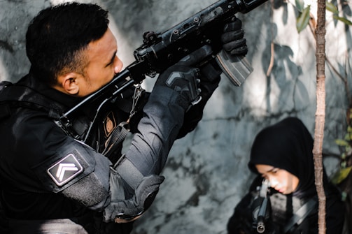 A person dressed in tactical gear aims a rifle upward while another person in a black hood is partially visible in the background. The setting appears to be an outdoor area with some foliage.