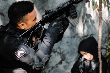 A person dressed in tactical gear aims a rifle upward while another person in a black hood is partially visible in the background. The setting appears to be an outdoor area with some foliage.