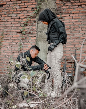 Two individuals dressed in tactical gear are positioned next to a brick wall with overgrown foliage. One person is crouched down, adjusting or checking equipment on the other's leg. Both are wearing black jackets and light-colored pants, with visible tactical vests.