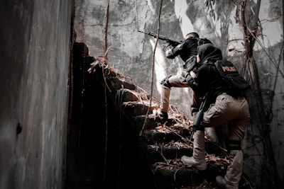 Two individuals in tactical police gear are cautiously ascending a set of overgrown concrete stairs in a dimly lit environment. They are armed and appear to be in a state of alertness, suggesting a potential mission or investigation in a derelict location.