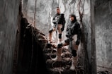 Two individuals in tactical gear stand confidently on a set of dilapidated stone stairs surrounded by a gloomy, overgrown building. They are holding rifles and wearing knee pads and gloves, exuding a sense of readiness and alertness. Sunlight filters through cracks, casting shadows on the walls.
