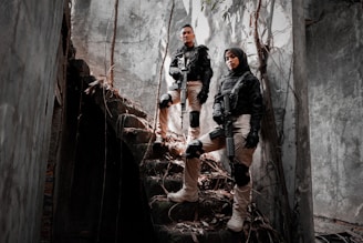 Two individuals in tactical gear stand confidently on a set of dilapidated stone stairs surrounded by a gloomy, overgrown building. They are holding rifles and wearing knee pads and gloves, exuding a sense of readiness and alertness. Sunlight filters through cracks, casting shadows on the walls.