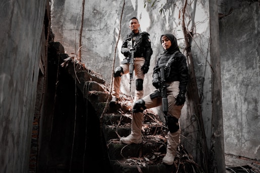 Two individuals in tactical gear stand confidently on a set of dilapidated stone stairs surrounded by a gloomy, overgrown building. They are holding rifles and wearing knee pads and gloves, exuding a sense of readiness and alertness. Sunlight filters through cracks, casting shadows on the walls.
