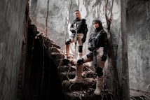 Two individuals in tactical gear stand confidently on a set of dilapidated stone stairs surrounded by a gloomy, overgrown building. They are holding rifles and wearing knee pads and gloves, exuding a sense of readiness and alertness. Sunlight filters through cracks, casting shadows on the walls.