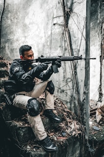 A person wearing tactical gear and knee pads is crouched on a rocky surface with dry leaves, holding a rifle. The background is a concrete wall with exposed roots and cracked surfaces, giving a sense of an abandoned or urban environment.