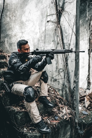 A person wearing tactical gear and knee pads is crouched on a rocky surface with dry leaves, holding a rifle. The background is a concrete wall with exposed roots and cracked surfaces, giving a sense of an abandoned or urban environment.