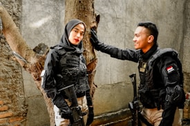 Two individuals in tactical gear stand by a tree against a wall with one person smiling at the other. Both wear black uniforms with tactical vests and hold rifles. The setting appears to be outdoor with a rough, textured wall and tree in the background.