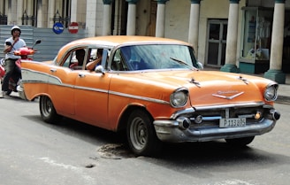 An orange vintage car is parked on a street with its doors slightly open. A man on a red scooter and a motorbike is seen nearby, wearing a helmet. The background features a building with columns, marble walls, and a shop with glass windows.