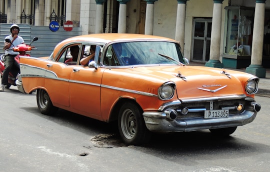 An orange vintage car is parked on a street with its doors slightly open. A man on a red scooter and a motorbike is seen nearby, wearing a helmet. The background features a building with columns, marble walls, and a shop with glass windows.