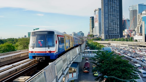 A modern train is moving along an elevated railway track above a bustling urban area. The background features a skyline dominated by tall skyscrapers and various buildings. Below the train tracks, a busy road is filled with numerous cars and buses. Lush green trees add contrast to the concrete structures.