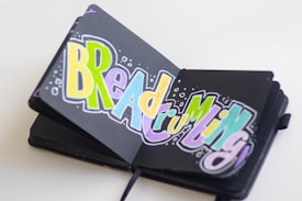 An open sketchbook displays colorful, bold and stylized lettering that spells 'Breadcrumbing' against a black background. The letters are filled with shades of yellow, blue, pink, and green, accentuated by dynamic white outlines and bubble-like embellishments.