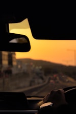 Close-up of a car steering wheel with a city street visible through the windshield during sunset.