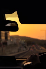 Close-up of a car steering wheel with a city street visible through the windshield during sunset.