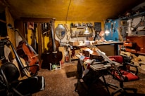 A cozy workshop with warm lighting where a person works at a table surrounded by various musical instruments like cellos and violins. The walls are lined with tools and equipment, and a mirror hangs amongst the clutter. The atmosphere suggests a focused and creative environment.