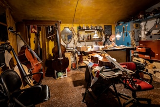 A cozy workshop with warm lighting where a person works at a table surrounded by various musical instruments like cellos and violins. The walls are lined with tools and equipment, and a mirror hangs amongst the clutter. The atmosphere suggests a focused and creative environment.