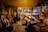 A cozy workshop with warm lighting where a person works at a table surrounded by various musical instruments like cellos and violins. The walls are lined with tools and equipment, and a mirror hangs amongst the clutter. The atmosphere suggests a focused and creative environment.