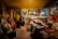 A cozy workshop with warm lighting where a person works at a table surrounded by various musical instruments like cellos and violins. The walls are lined with tools and equipment, and a mirror hangs amongst the clutter. The atmosphere suggests a focused and creative environment.