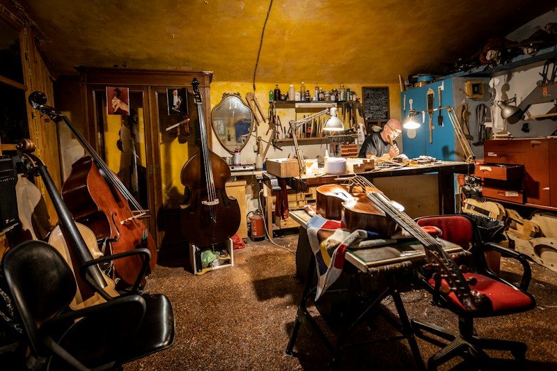 A cozy workshop with warm lighting where a person works at a table surrounded by various musical instruments like cellos and violins. The walls are lined with tools and equipment, and a mirror hangs amongst the clutter. The atmosphere suggests a focused and creative environment.