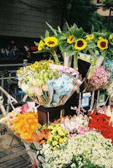 A diverse assortment of flowers is displayed in bouquets at an outdoor flower stand. Sunflowers, roses, daisies, and other flowers in a range of colors including yellow, pink, blue, red, and white are visible. There are also price tags and QR codes attached to several bouquets. In the background, bicycles and a street can be seen.