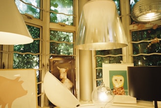 A cozy and artistic interior setting featuring multiple lampshades and framed art. The background showcases a large window with green foliage visible through the glass. The artwork includes a framed picture of a sheep and an owl. The soft light and decorative items create a warm and inviting atmosphere.
