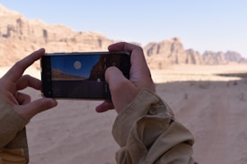 A person wearing a beige jacket is holding a smartphone, capturing a photo of a desert landscape with rocky formations in the background. The scene includes sand dunes under a clear blue sky, visible both in the foreground and on the smartphone screen.