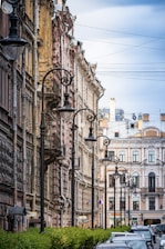 A narrow urban street lined with historic buildings featuring ornate architectural details. Tall, black street lamps are aligned in a row alongside the buildings, and the street is bordered by green hedges. There are a few cars parked along the street, and the sky is partially cloudy.