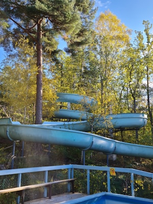 A winding water slide surrounded by lush greenery and trees in a natural setting. The structure is a light blue color, contrasting with the vibrant yellow and green leaves. Sunlight filters through the tall trees, creating a warm and inviting atmosphere.