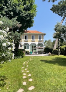 A charming two-story house with a yellow facade and a red-tiled roof is surrounded by lush greenery. The front yard features a neatly maintained lawn with a stone path leading to the patio. There are outdoor furniture pieces, such as chairs and a table under a large umbrella.