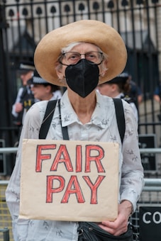 An older person wearing a straw hat, eyeglasses, and a face mask stands holding a cardboard sign with the words 'FAIR PAY' written in bold red letters. There is a subtle pattern on their light-colored shirt, and a couple of police officers are visible in the background against a black iron fence.