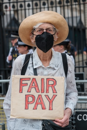 An older person wearing a straw hat, eyeglasses, and a face mask stands holding a cardboard sign with the words 'FAIR PAY' written in bold red letters. There is a subtle pattern on their light-colored shirt, and a couple of police officers are visible in the background against a black iron fence.