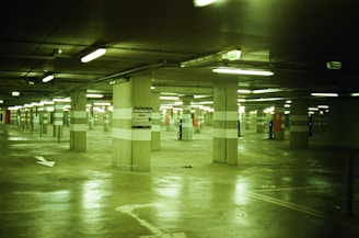 A dimly lit underground parking garage with concrete pillars and fluorescent lighting. The floor is reflective, and there are marked parking spaces with some signs visible on the pillars.