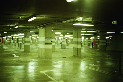 A dimly lit underground parking garage with concrete pillars and fluorescent lighting. The floor is reflective, and there are marked parking spaces with some signs visible on the pillars.