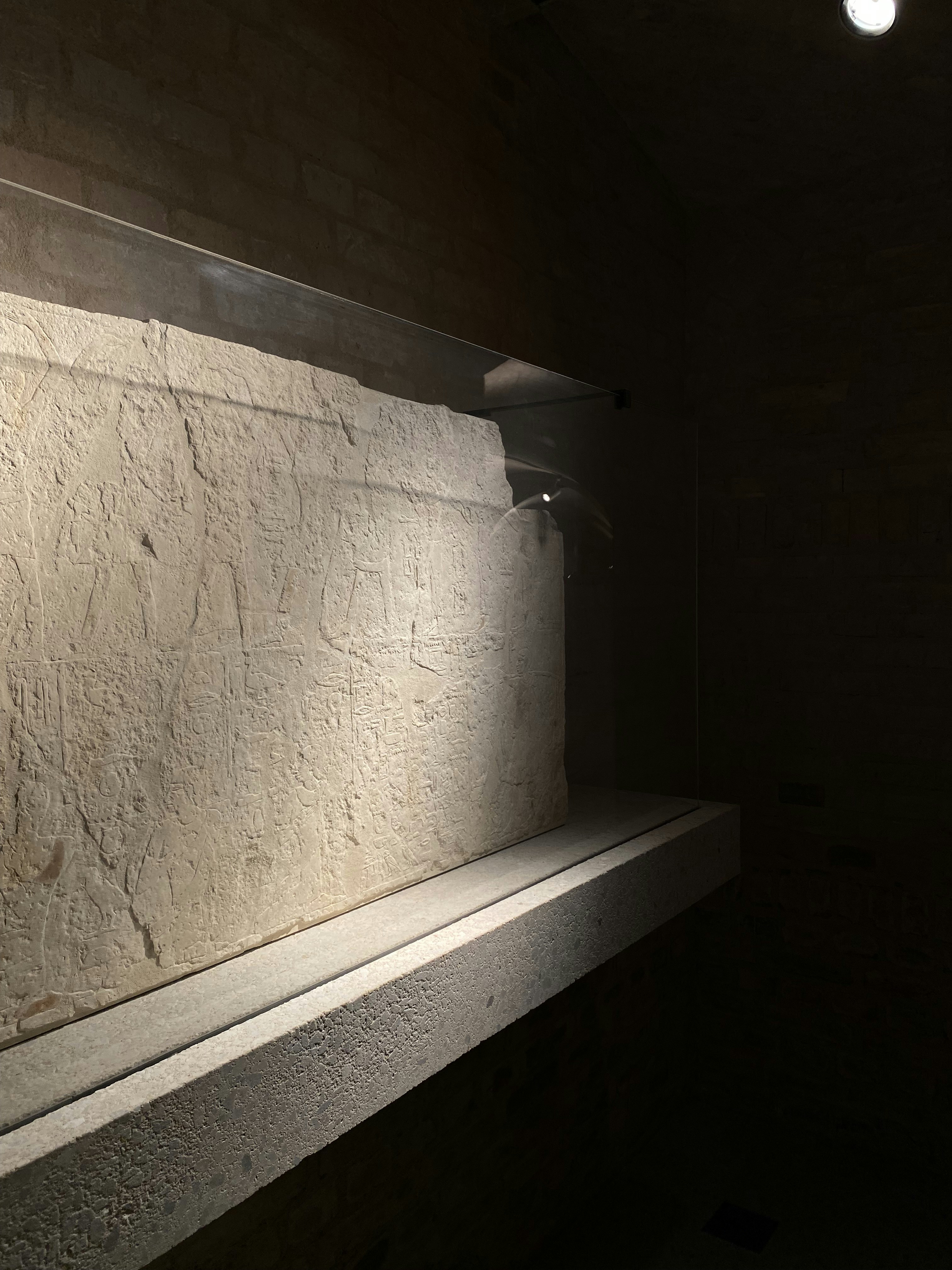 Ancient stone inscription tablet in dim museum light, carved glyphs visible, deep shadows, dramatic spotlight, dark stone walls