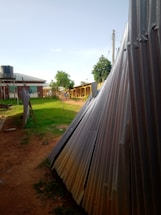 Corrugated metal sheets are stacked beside a path leading to a row of buildings with a water tank on stilts nearby. There are a few small trees and grass lawns along the path, and power lines are visible above. The scene is lit by natural daylight under a clear sky.